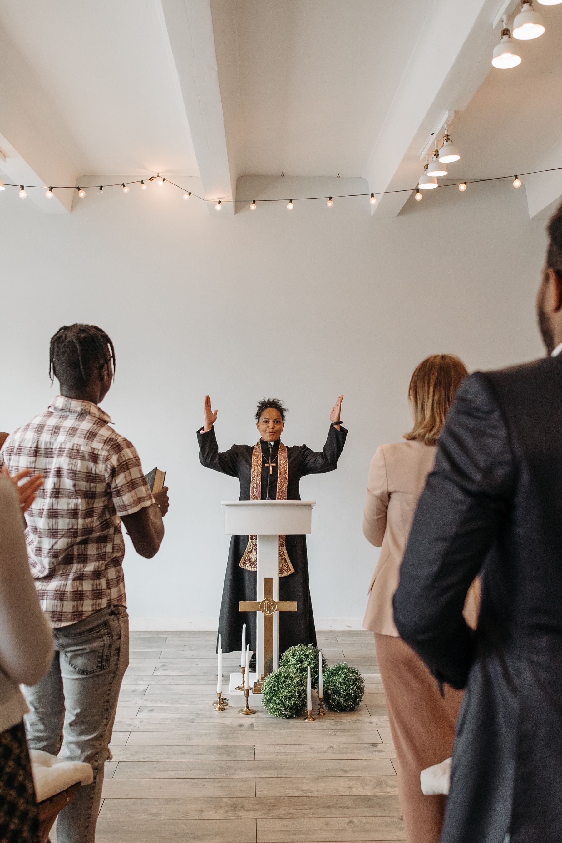 A pastor at a white podium with hands raised as people stand